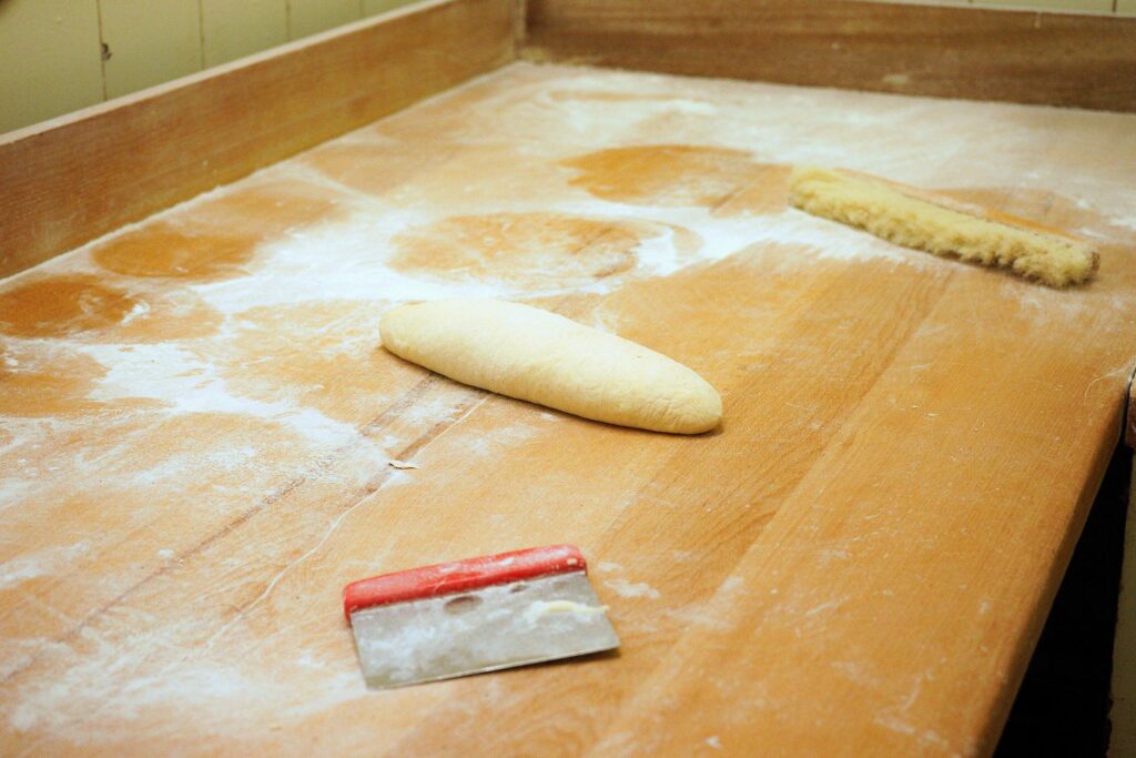 Ein Baguette Teigrohling auf einer Holzarbeitsfläche davor ein Stechspachtel und im Hintergrund ein Tischbesen.